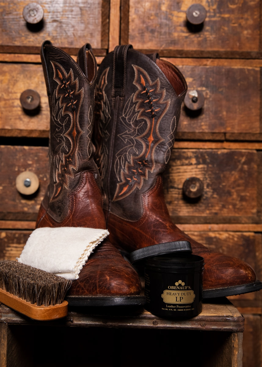 A pair of brown leather cowboy boots with carved western embroidery, displayed on a wooden surface with a white cloth, a brush, and a black container of O'Neal's Leather Care LP against a rustic wooden drawer background.
