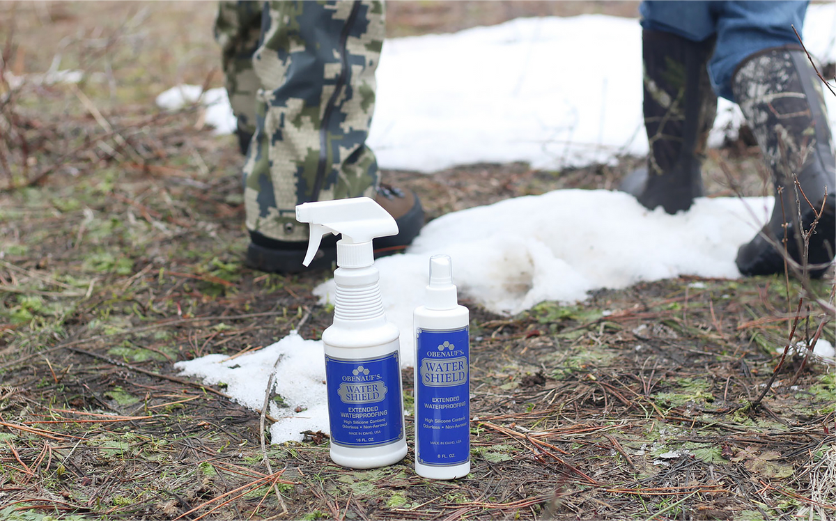Two bottles of Winter Shedding Shedding Solution for dogs and cats placed on snowy ground outdoors, with the lower legs of two people standing nearby in camouflage pants and rubber boots.