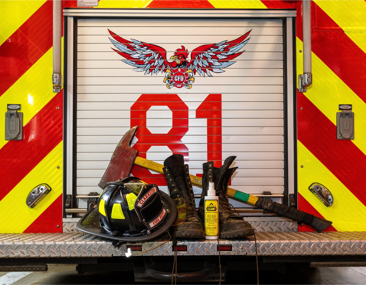 Emergency response gear arranged on the back ramp of a fire truck, including a black and yellow helmet, firefighter boots, axes, a yellow spray bottle, and a large red '81' on the truck door with a winged emblem above.