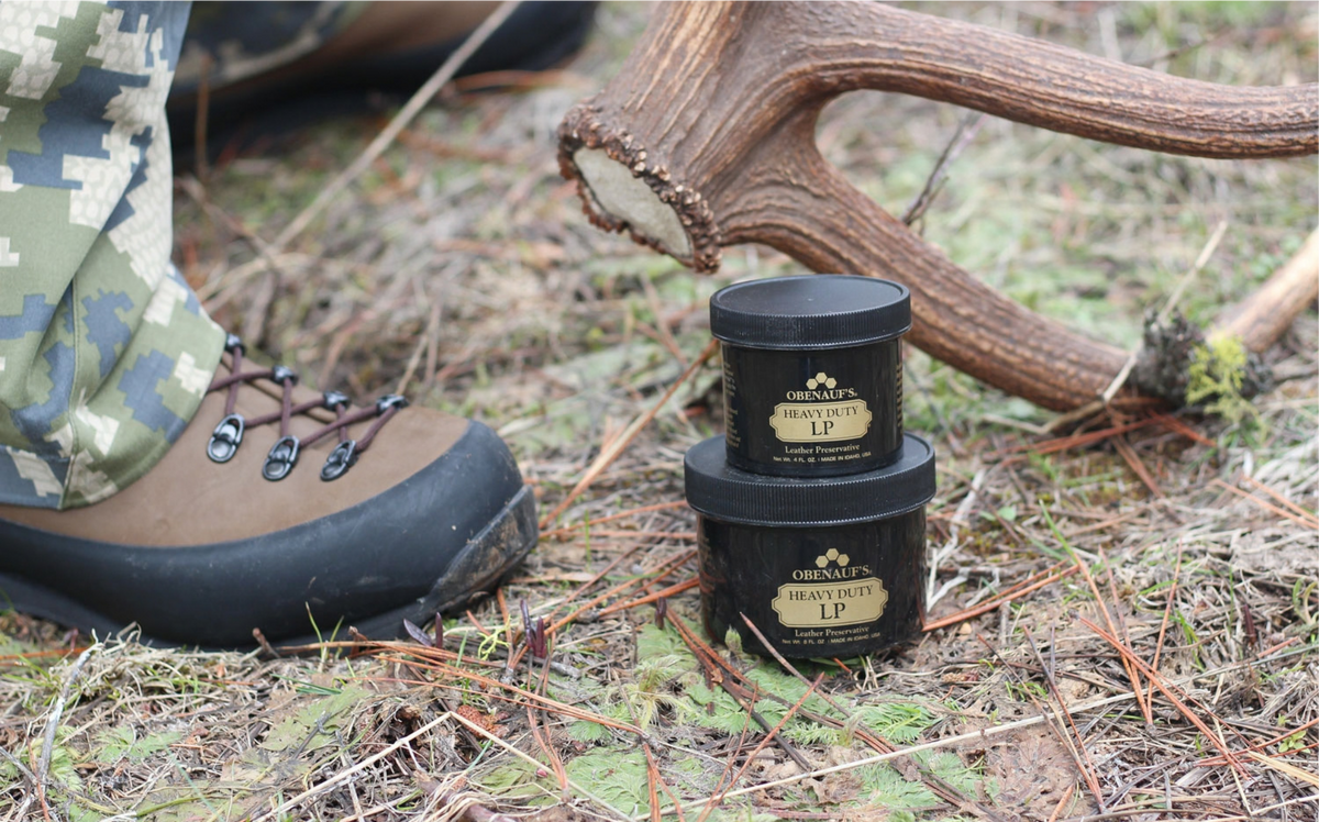 Two black cosmetic jars of Groomer's Lip Balm stacked on the ground outdoors, with a person's boot and camouflage pants visible on the left and a deer antler in the background.
