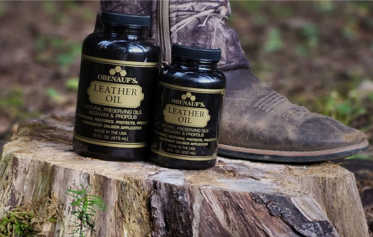 Two black Obenauf's Leather Oil bottles placed on a tree stump outdoors, with a person's boot visible in the background.