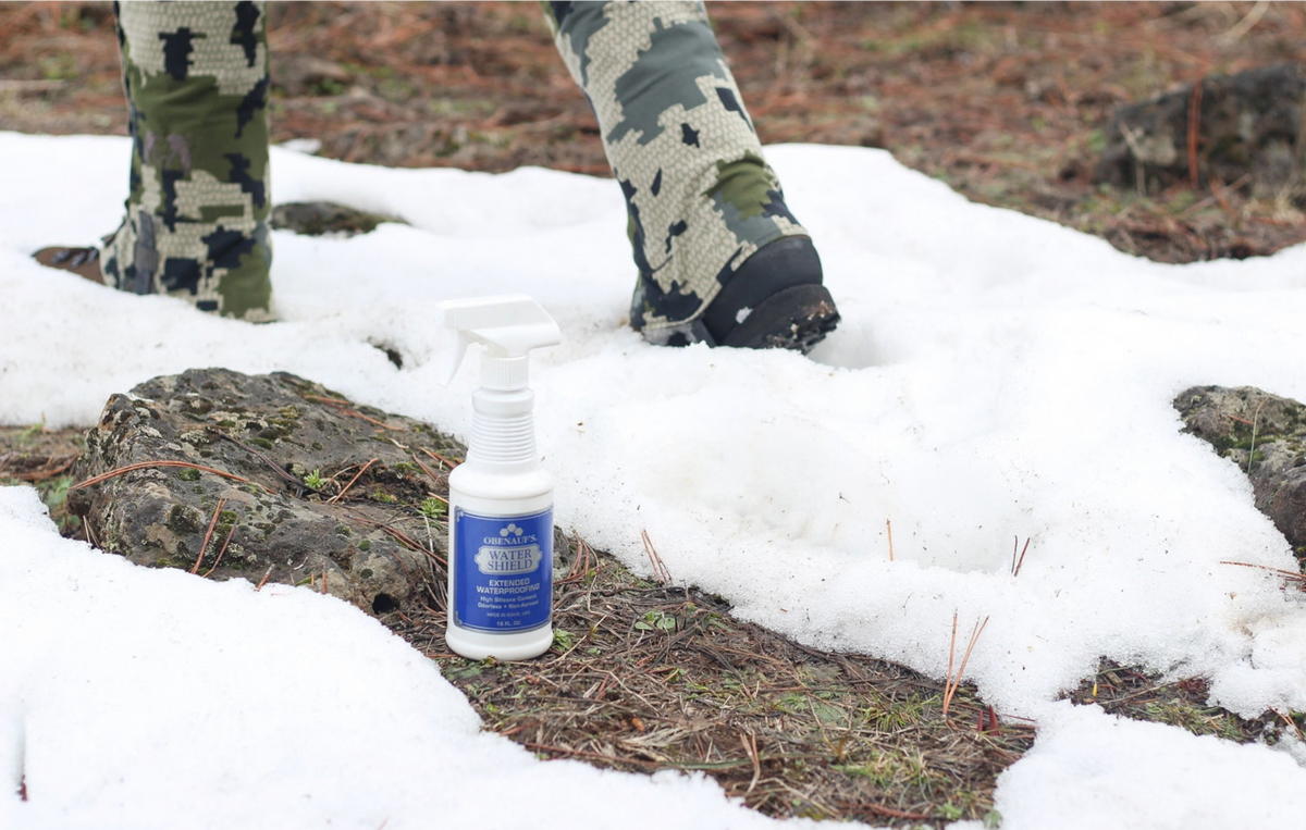 A white Water Shield spray bottle with a blue label labeled 'STAIN REMOVER' is placed on a snowy forest ground next to a person's legs wearing camouflage pants.