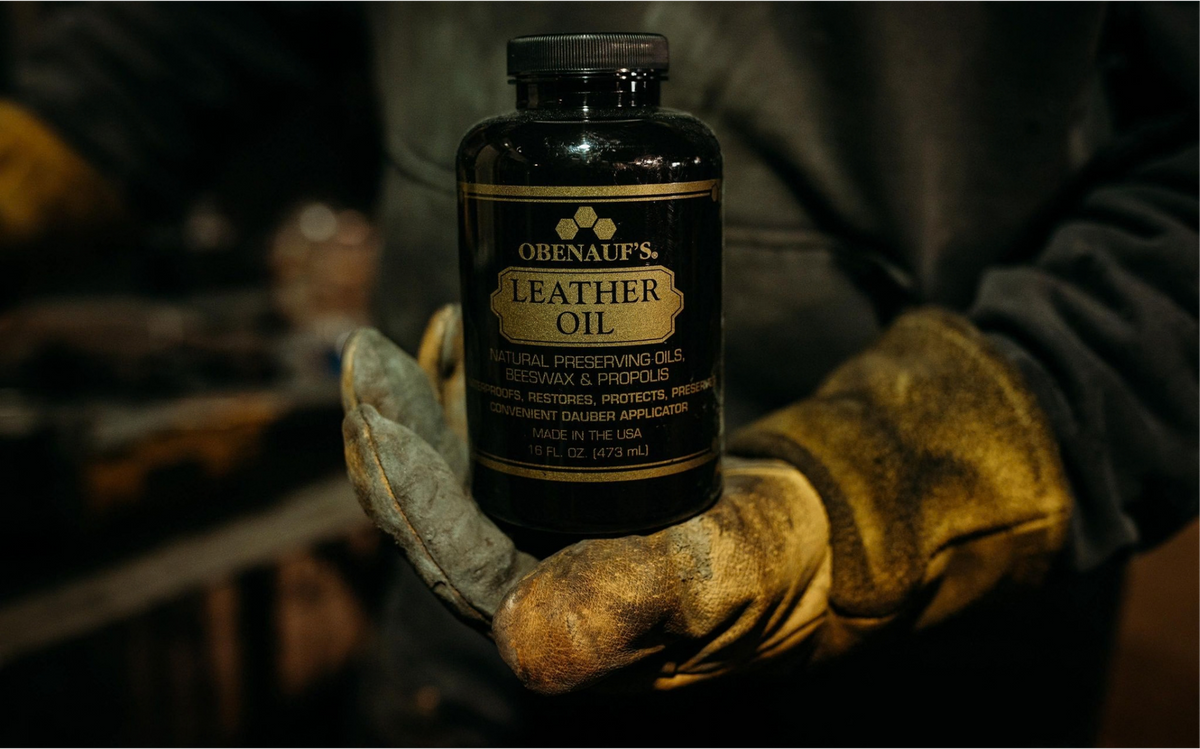 A person wearing heavy-duty leather oil gloves holds a bottle of Obenauf's Leather Oil in a dark workshop setting.