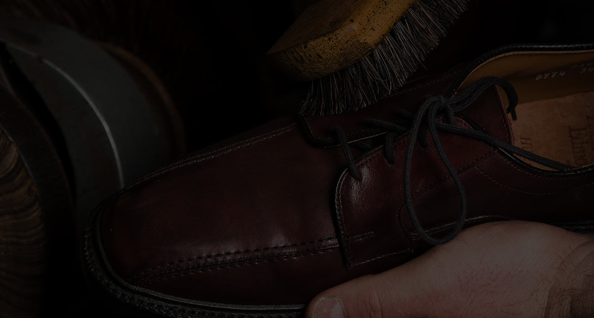 A close-up of a dark brown leather lace-up shoe held by a hand, with a wooden brush visible in the background against a dark setting.